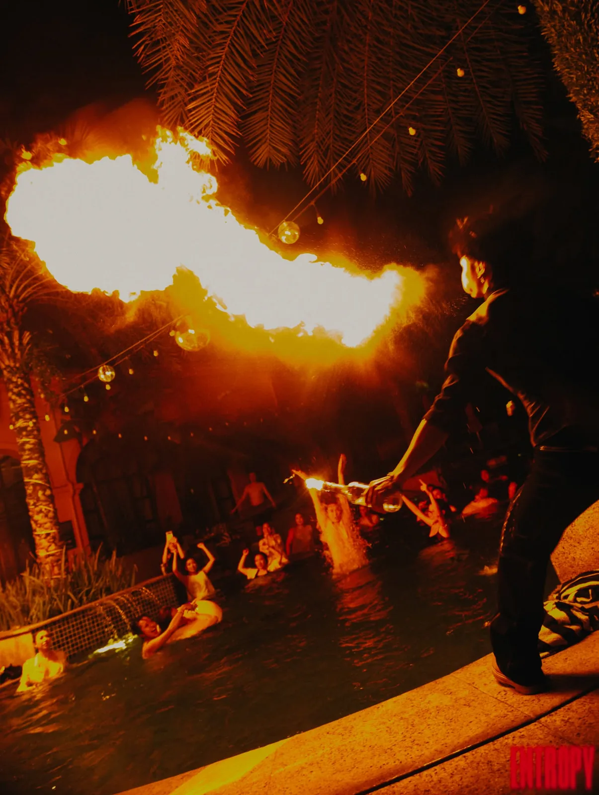 A fire performer breathing flame over a lit pool at night